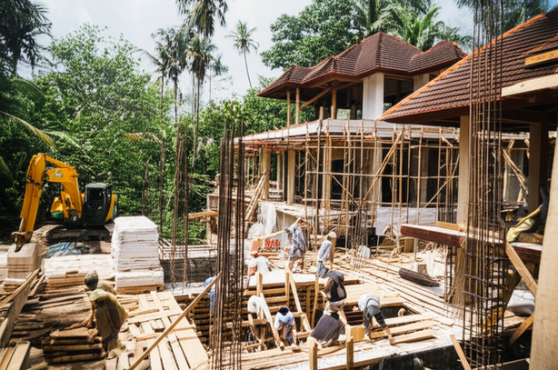 Construction of a Bali villa, showing various stages from foundation work to structural framework, surrounded by lush tropical vegetation.