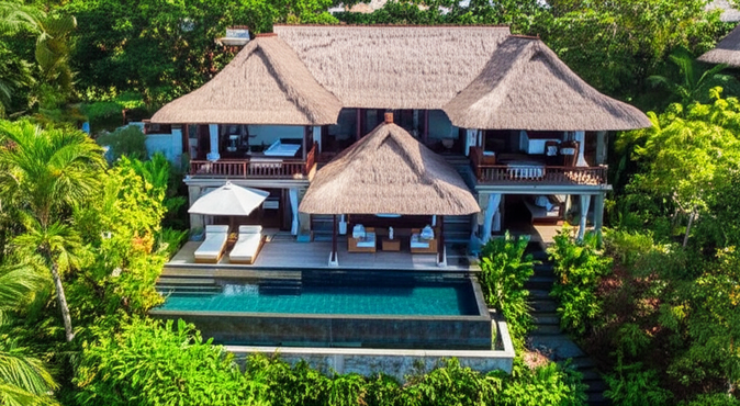 Aerial view of a Balinese villa showcasing traditional architecture integrated with the natural landscape, featuring thatched roofs, an infinity pool, and tropical gardens.
