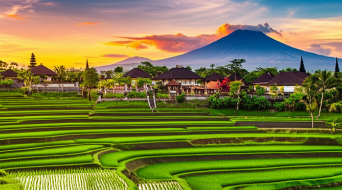 Panoramic view of Balinese landscape, showcasing terraced rice paddies, traditional buildings, and volcanic mountains at sunset, illustrating Bali's building height restrictions.